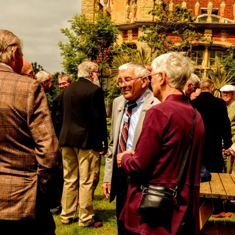 A group of older adults dressed in formal attire converse outdoors in a garden near a brick building, with a wooden picnic table visible in the foreground.