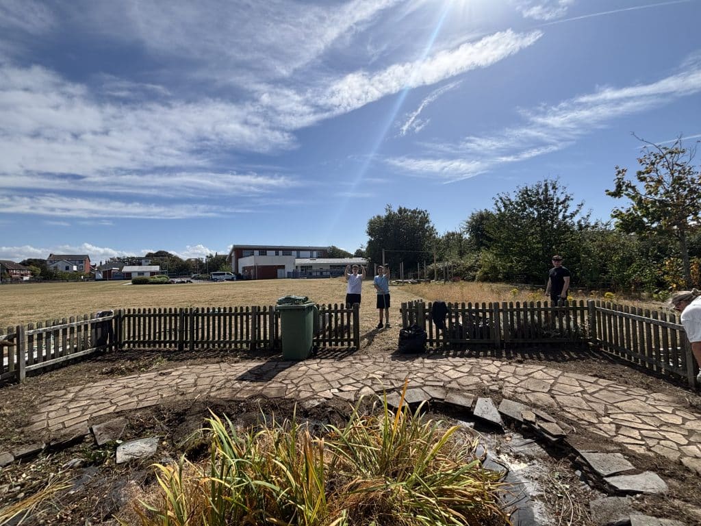 A small fenced garden with two people standing behind the fence, one person to the right, under a mostly sunny sky with houses and trees in the background.