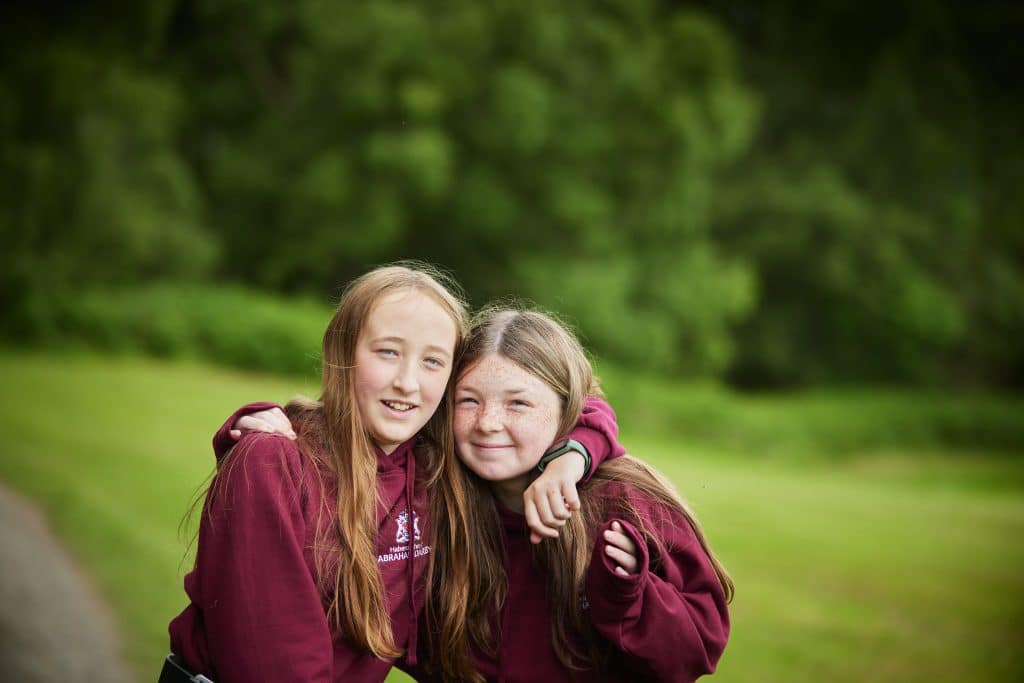 Two girls in matching maroon hoodies stand outdoors with their arms around each other, smiling at the camera. Green trees and grass are in the background.
