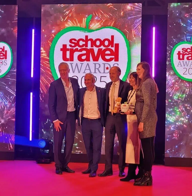 Five people stand on a red carpet in front of a "School Travel Awards 2023" display, posing for a photo with an award and a teddy bear.