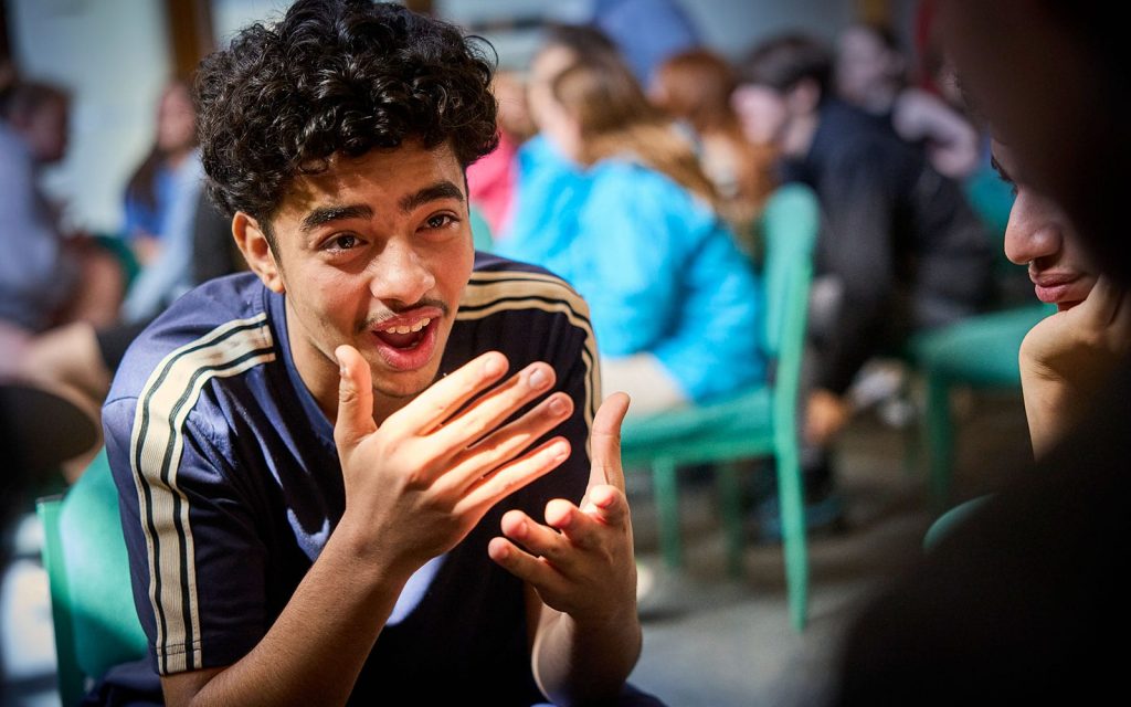 A young person with curly hair gestures while speaking, sitting in a room with others who are blurred in the background.