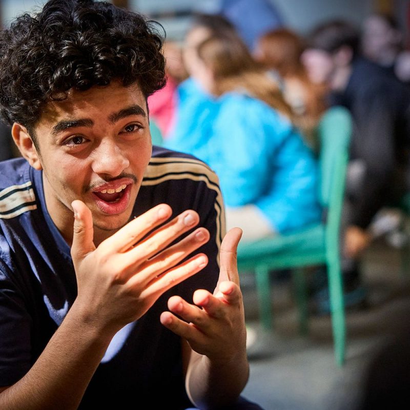 A young person with curly hair gestures while speaking, sitting in a room with others who are blurred in the background.