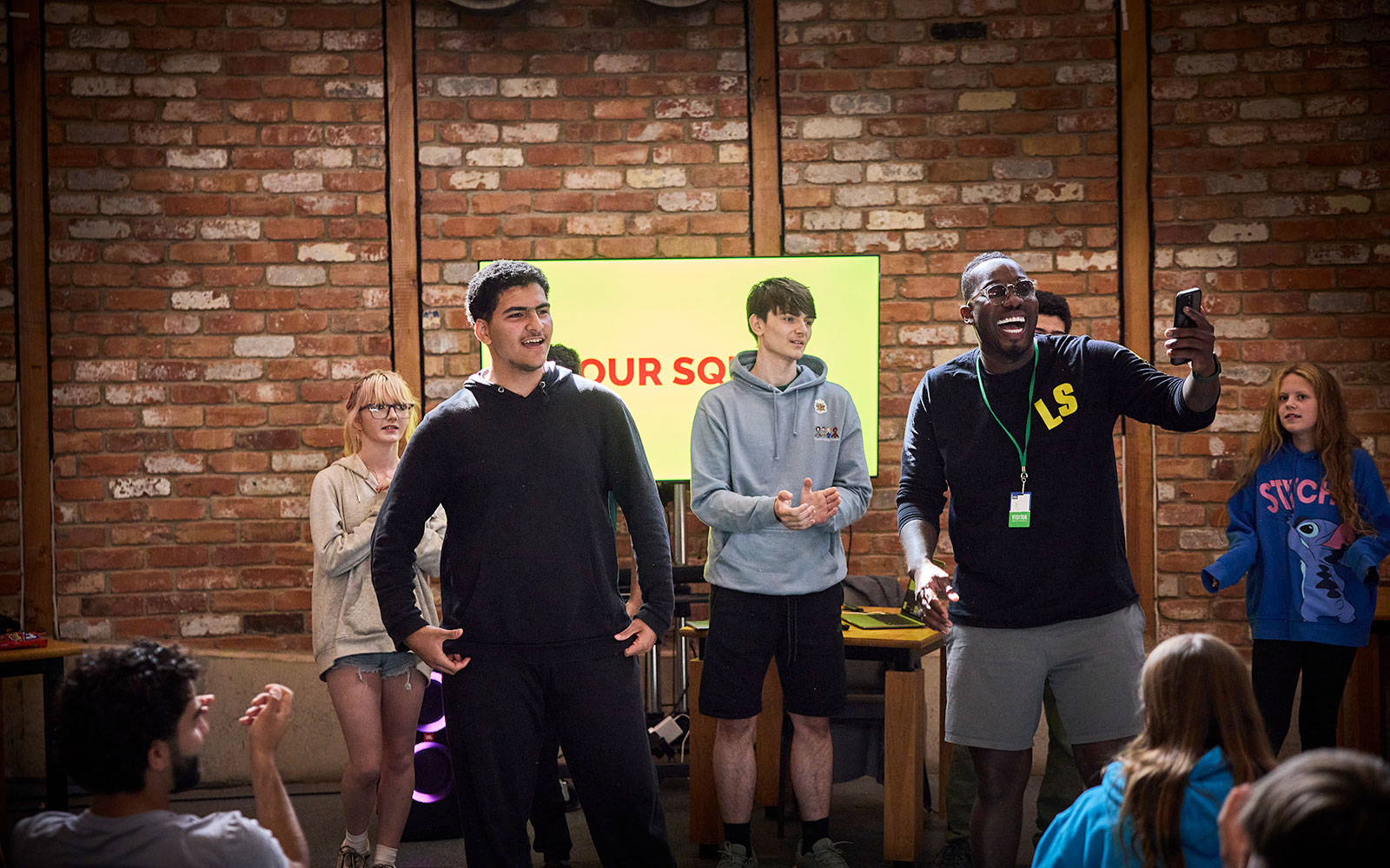 A group of young adults stands in front of a brick wall, some clapping and smiling, while one person takes a selfie with others in the background.
