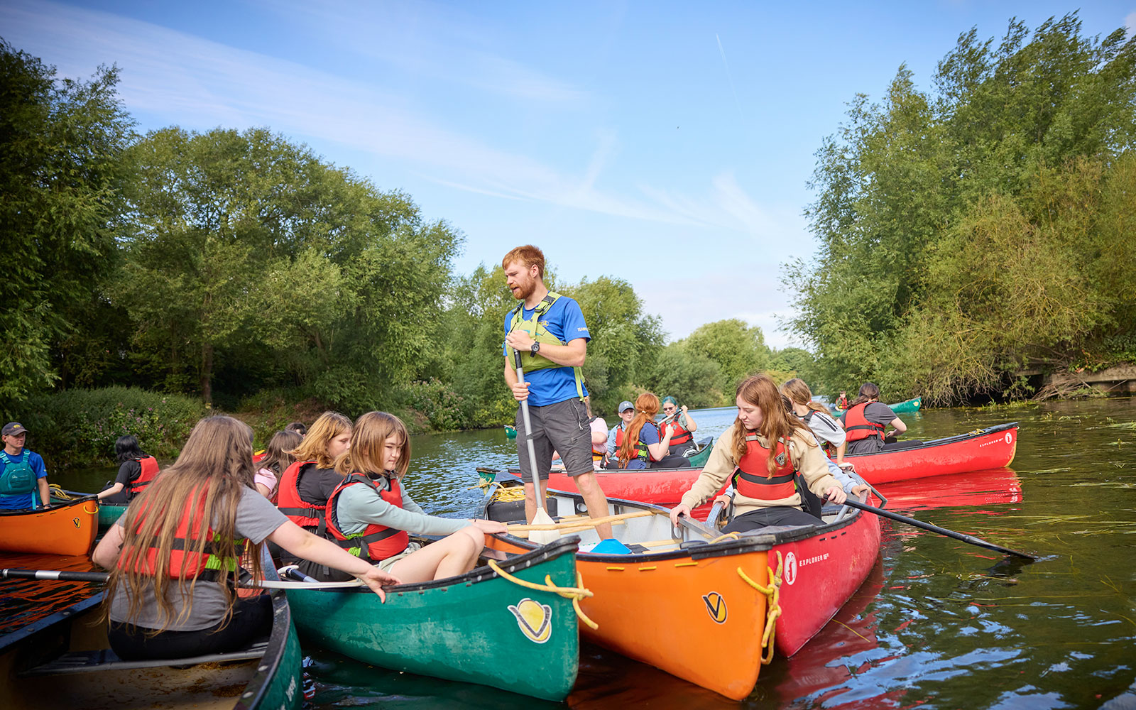 A group of people in life jackets sit in colorful canoes on a calm river, with an instructor standing and giving guidance. Trees and blue sky are visible in the background.