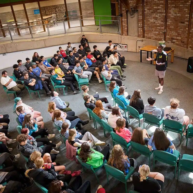 A group of people seated in an indoor amphitheater listen to a person speaking at the front near a yellow screen.
