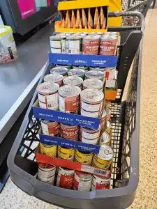 A shopping basket filled with stacked canned foods, including Irish stew, minced beef and onion, and spaghetti bolognese, next to a box of instant noodle packets at a checkout.