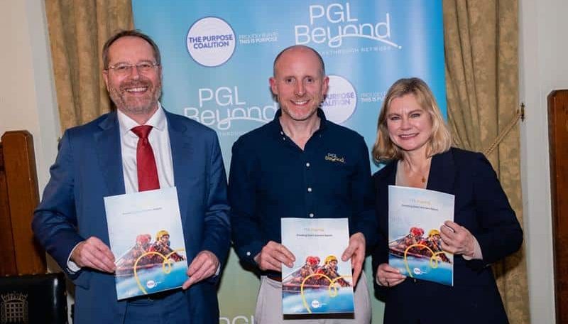 Three people stand indoors holding up booklets in front of a PGL Beyond banner, smiling for the camera.