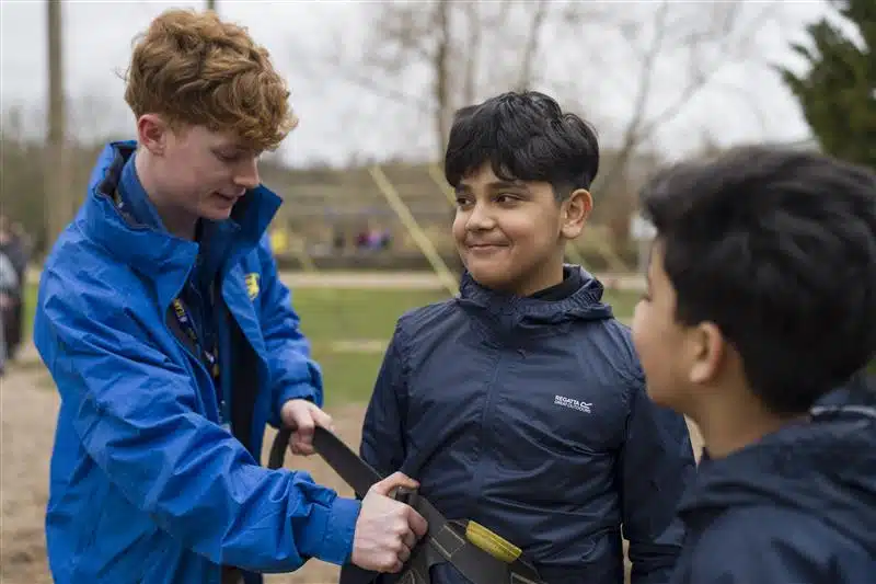 A young instructor helps a boy fasten a safety harness outdoors whilst another boy stands nearby.