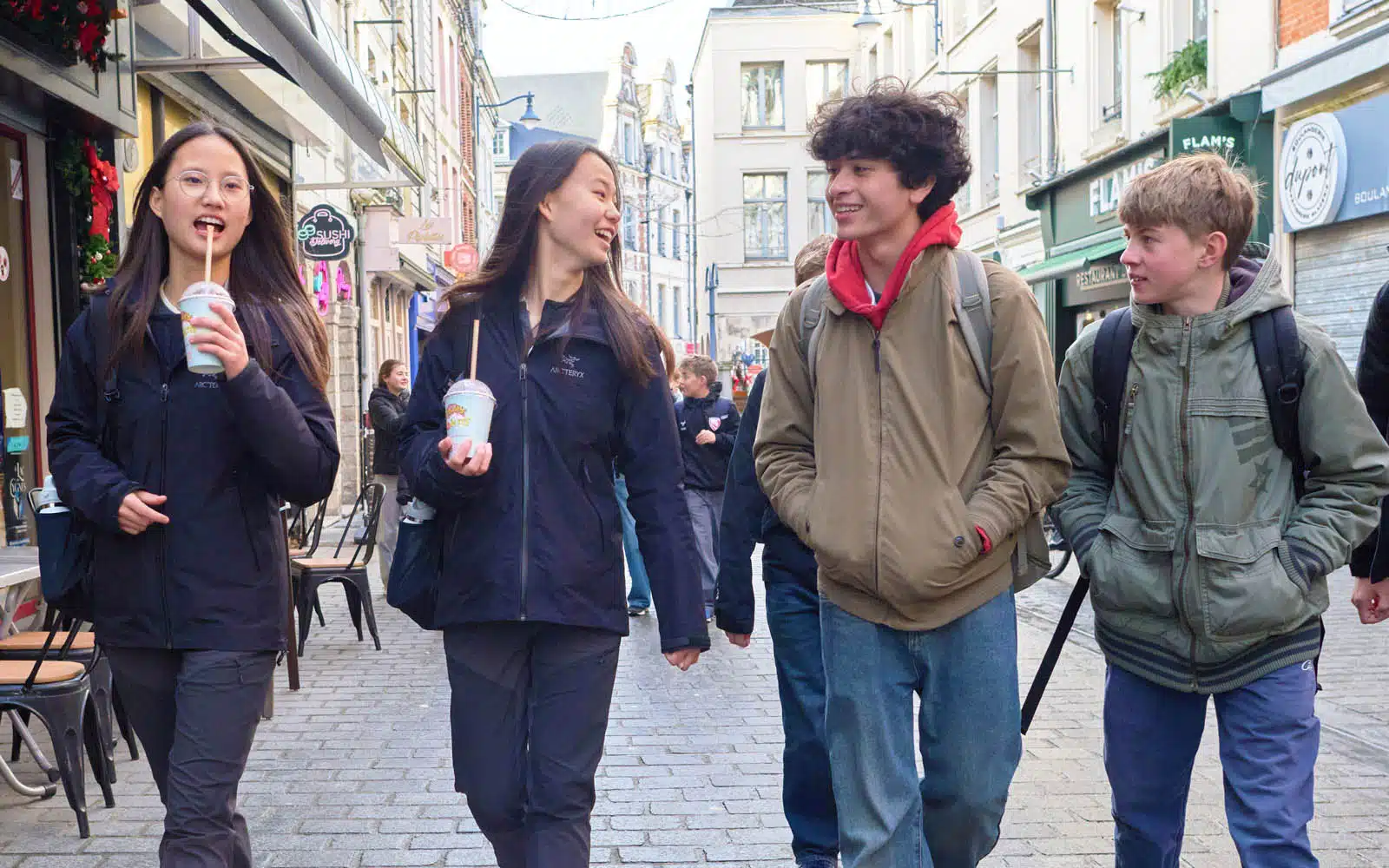 Four teenagers walk down a city street, chatting and drinking beverages. They wear jackets and rucksacks, and there are shops and pedestrians in the background.