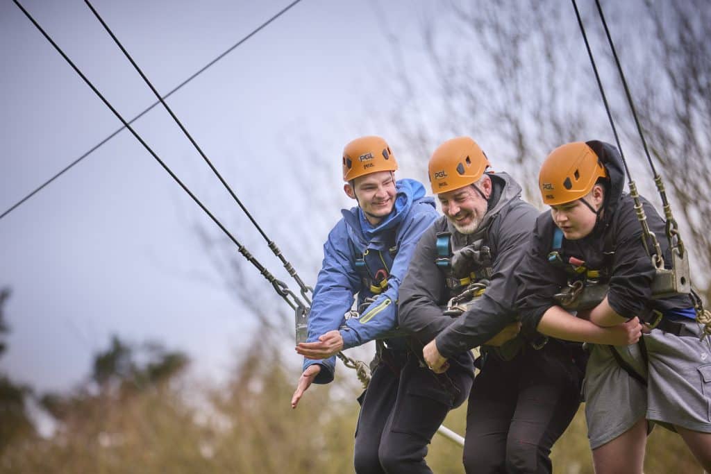 Three people wearing helmets and harnesses are suspended on a zip wire outdoors, preparing to launch together. Trees are visible in the background.