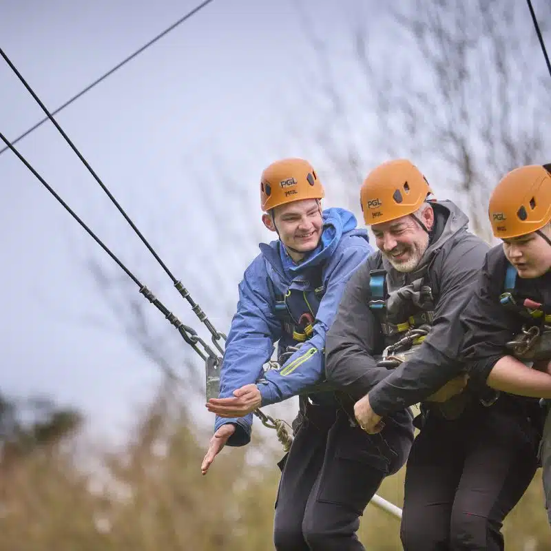 Three people wearing helmets and harnesses are suspended on a zip wire outdoors, preparing to launch together. Trees are visible in the background.