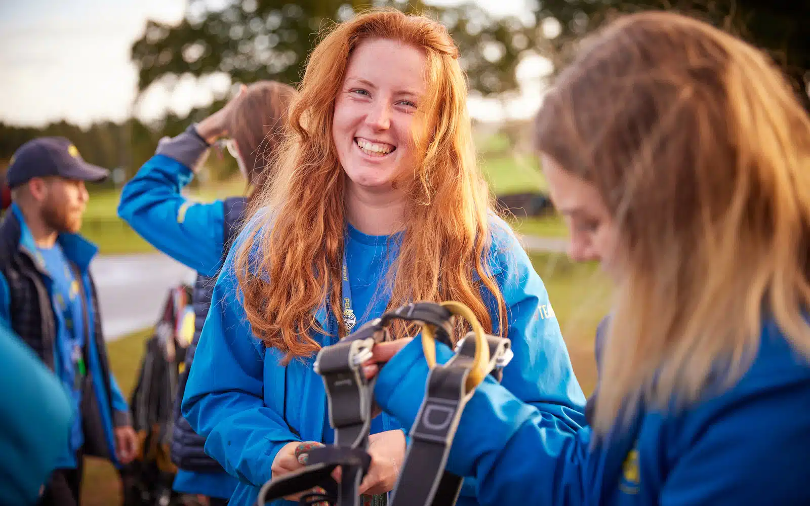 A woman with long red hair and a blue jacket smiles outdoors whilst another person holds climbing harnesses in the foreground. Other people are visible in the background.