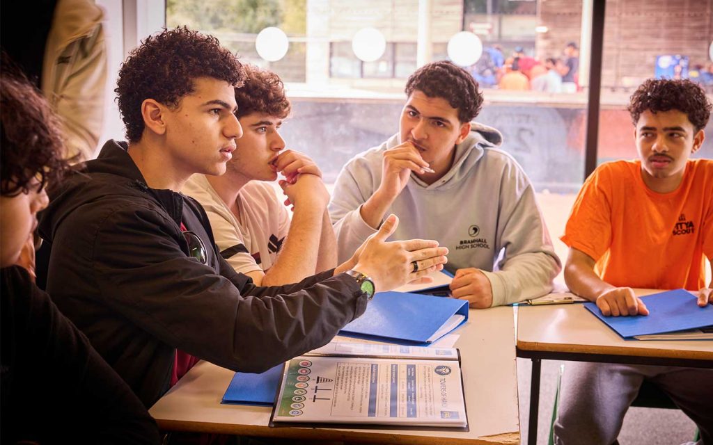 Four young men sit at a table with blue folders, engaged in discussion, whilst one gestures with his hand and the others listen attentively.