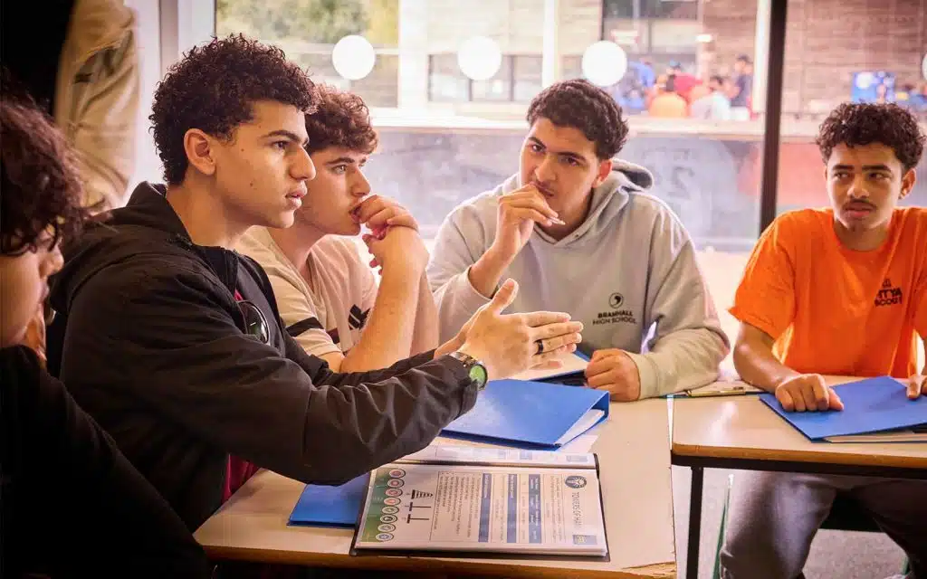 Four young men sit at a table with blue folders, engaged in discussion, whilst one gestures with his hand and the others listen attentively.
