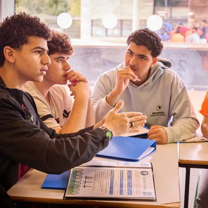 Four young men sit at a table with blue folders, engaged in discussion, whilst one gestures with his hand and the others listen attentively.
