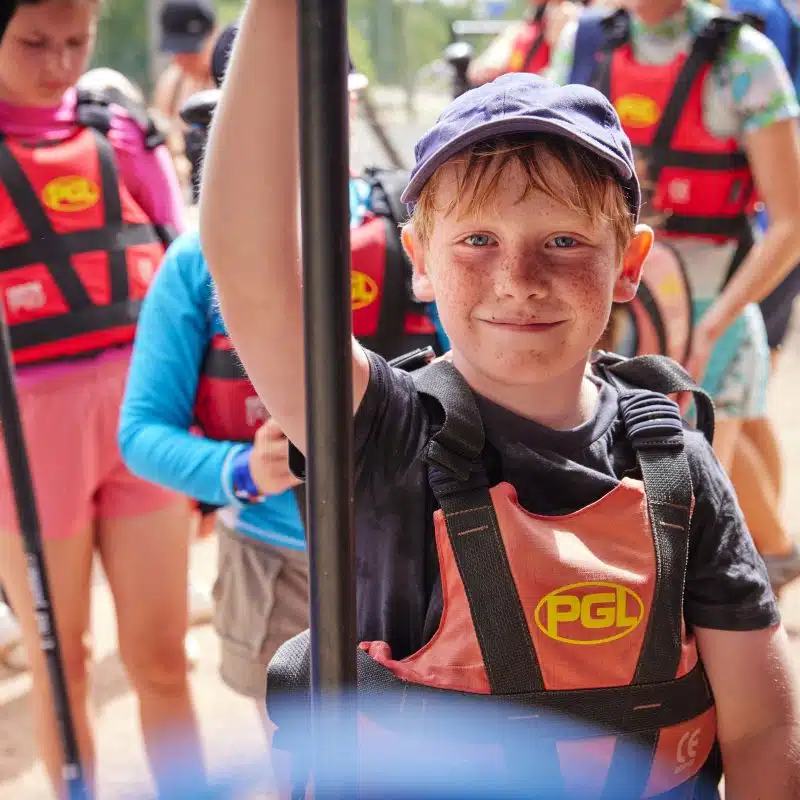 A boy wearing a blue cap and a life jacket holds a paddle, standing among other children in life jackets, possibly at an outdoor camp or water activity.