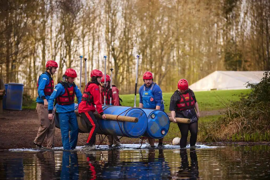 Five people in helmets and life jackets carry a homemade raft made from blue barrels and wooden poles into a lake, with trees and grass in the background.