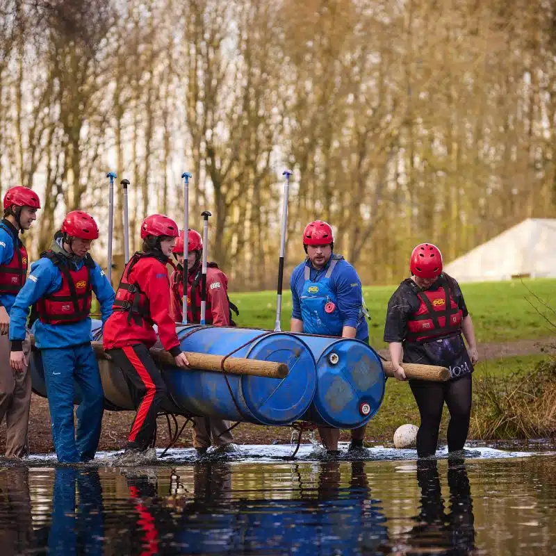 Five people in helmets and life jackets carry a homemade raft made from blue barrels and wooden poles into a lake, with trees and grass in the background.