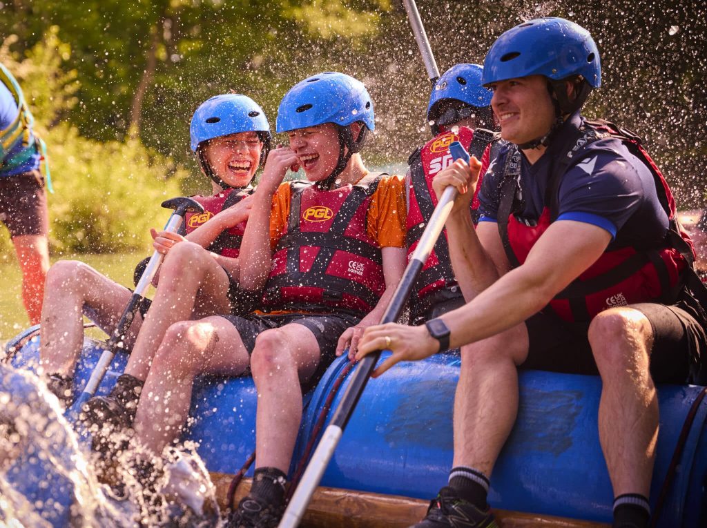 Four people in blue helmets and life jackets laughing while white-water rafting and paddling through a splash of water.